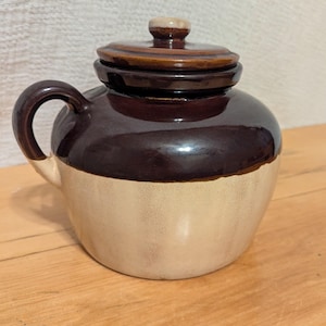 Puede incluir: Un tarro de cerámica vintage de dos tonos con tapa. El tarro es de color marrón oscuro en la parte superior y beige claro en la inferior. Tiene un asa curva y una tapa marrón a juego con un pequeño pomo. El tarro está sobre una superficie de madera.