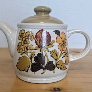 Puede incluir: Tetera de cerámica color crema con tapa y asa. La tetera está decorada con motivos florales en tonos amarillos, marrones y rojos. El pico y el asa son blancos, y la tapa es de color marrón claro.