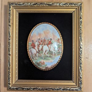 Könnte beinhalten: Ein ovales, gerahmtes Kunstwerk, das eine Jagdszene mit Reitern auf weißen Pferden zeigt, die rote Mäntel und schwarze Zylinder tragen. Das Kunstwerk ist vor einem schwarzen Hintergrund in einem goldfarbenen, verzierten Rahmen angeordnet. Die Szene umfasst einen Fluss und Bäume.