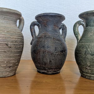 Puede incluir: Tres jarrones antiguos de cerámica con asas. El jarrón de la izquierda es de color gris claro con líneas horizontales. El jarrón del medio es de color marrón oscuro y el de la derecha es verde. Los tres jarrones tienen un aspecto envejecido.
