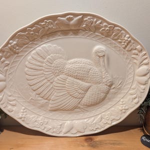 Peut inclure: Un plat ovale en céramique de couleur crème avec un relief représentant une dinde au centre. La bordure est ornée de fruits, de feuilles et d'un motif en treillis en relief. La surface présente une finition brillante et un bord festonné.