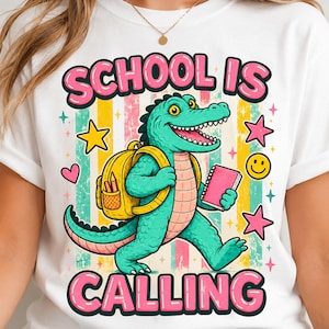 Puede incluir: Una camiseta blanca con un gráfico de un caimán verde que lleva una mochila amarilla y sostiene un cuaderno rosa. El texto "SCHOOL IS CALLING" aparece en letras de burbuja rosas arriba y abajo del caimán, rodeado de estrellas y corazones.