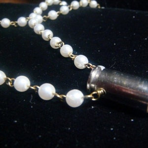 Può includere: Una collana con piccole perle bianche collegate da maglie color oro. Presenta un ciondolo a proiettile color argento con una perla bianca sulla punta. Il gioiello è esposto su uno sfondo scuro.