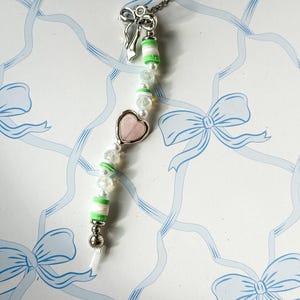 May include: A silver chain necklace with a green and white striped bead charm, a pink heart charm, and a silver bow charm. The necklace is on a white background with a blue ribbon pattern.
