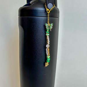 May include: A black water bottle with a gold chain and a beaded charm. The charm features a green and gold butterfly, white beads, and green and gold beads. The charm is attached to the bottle's handle.