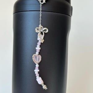 May include: A black water bottle with a decorative charm. The charm features a silver bow, pink heart-shaped bead, and various pink and white beads. The charm is attached to the bottle with a silver chain and clasp.