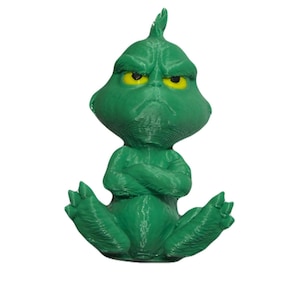 May include: A green 3D printed figurine of a creature with a large head, pointed hair, and a grumpy facial expression. The figure has yellow eyes, a furrowed brow, and arms crossed over its chest while sitting with legs extended outward.