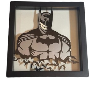 May include: A framed Batman art piece. The artwork features a gray and black silhouette of Batman, with his iconic bat symbol on his chest. The frame is black, and the background is a light gray. The bottom of the artwork has a bat-shaped design.
