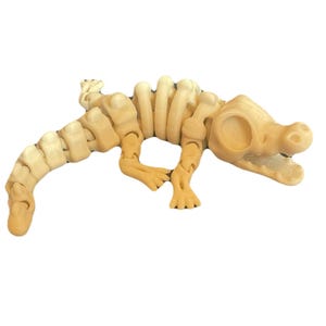 May include: A light brown articulated alligator skeleton toy. The toy is made of a light brown material and features a detailed skeletal structure, including ribs, vertebrae, and a skull. The alligator is posed on a white background.