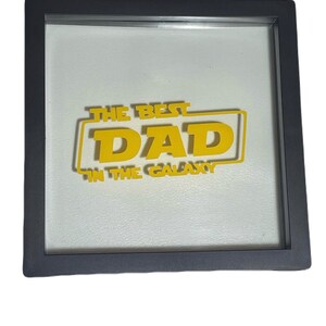 May include: A framed artwork with the text "THE BEST DAD IN THE GALAXY" in yellow, set against a white background. The text is enclosed in a yellow rectangular border, and the frame is dark gray.