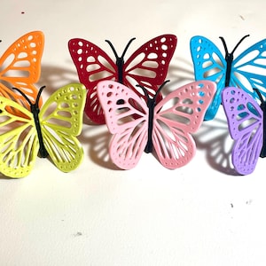 May include: A collection of colorful butterfly decorations in orange, red, blue, pink, purple, and yellow-green. Each butterfly has a detailed wing design and black body. These decorative butterflies are suitable for crafts and home decor.