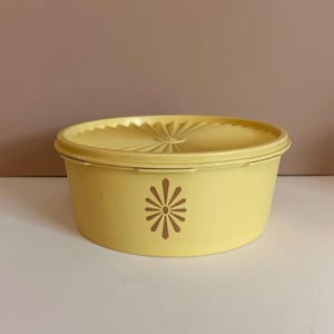 Puede incluir: Un recipiente de plástico amarillo vintage con tapa a juego. La tapa tiene un diseño de rayos de sol y el recipiente presenta un gráfico floral marrón. El recipiente es redondo y parece estar en buenas condiciones.