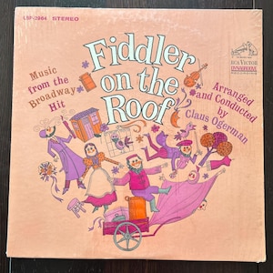 Może przedstawiać: Okładka winylowej płyty z muzyką z musicalu "Skrzypek na dachu". Okładka przedstawia kolorową ilustrację postaci z musicalu, w tym skrzypka, kobietę w długiej sukni i mężczyznę w kapeluszu. Tytuł "Skrzypek na dachu" jest wydrukowany dużymi, pogrubionymi literami na okładce.