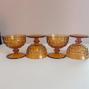 May include: Four amber-colored, vintage-style dessert or parfait glasses. Each glass features a round bowl with a textured pattern, supported by a short stem and a circular base. The glasses are arranged in a row, showcasing their design.