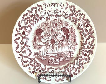 May include: A white ceramic Christmas plate with a maroon illustration. The plate features a family decorating a Christmas tree, surrounded by holly and candy cane designs. The words "Merry Christmas" and the year "1996" are also displayed.