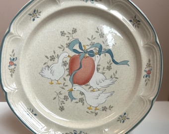 May include: A large, cream-colored ceramic dinner plate with a blue rim and a floral pattern. The center of the plate features a hand-painted design of three white geese with blue ribbons around a red apple.