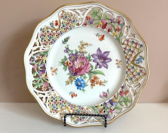 Peut inclure: Une assiette décorative en porcelaine blanche avec un bord festonné et une bordure dorée. L'assiette présente un arrangement floral central en rose, violet, bleu et orange. Le bord extérieur a un design ajouré avec des accents floraux colorés.