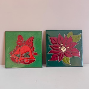 Puede incluir: Dos baldosas de cerámica cuadradas con diseños navideños. Una presenta campanas rojas con un lazo sobre un fondo verde. La otra baldosa muestra una flor de pascua roja con hojas verdes sobre un fondo verde. Ambas baldosas tienen un acabado brillante.