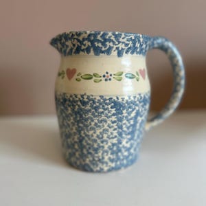 Puede incluir: Una jarra de cerámica azul y blanca con un diseño floral y corazones rosas. La jarra tiene un esmalte moteado y un cuerpo redondeado.