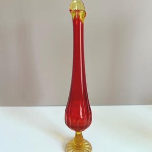 Puede incluir: Un jarrón de cristal rojo, alto y delgado, con borde y base amarillos. El jarrón tiene forma de lágrima y superficie texturizada. La base es redonda con diseño festoneado. Una pieza decorativa para el hogar.