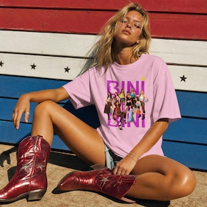 May include: A light pink short-sleeve t-shirt featuring a graphic print of a group of people and the text BINI in large purple letters. The shirt is paired with metallic red cowboy boots that have a star design on the side of the shaft.