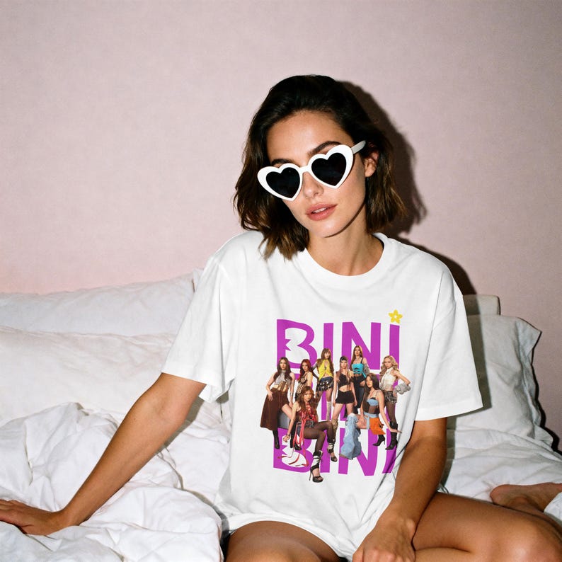 May include: A white short-sleeved t-shirt featuring a graphic print of a group of eight people in various outfits. The text BINI is printed in large purple letters behind the group, with a small yellow star icon above the text.