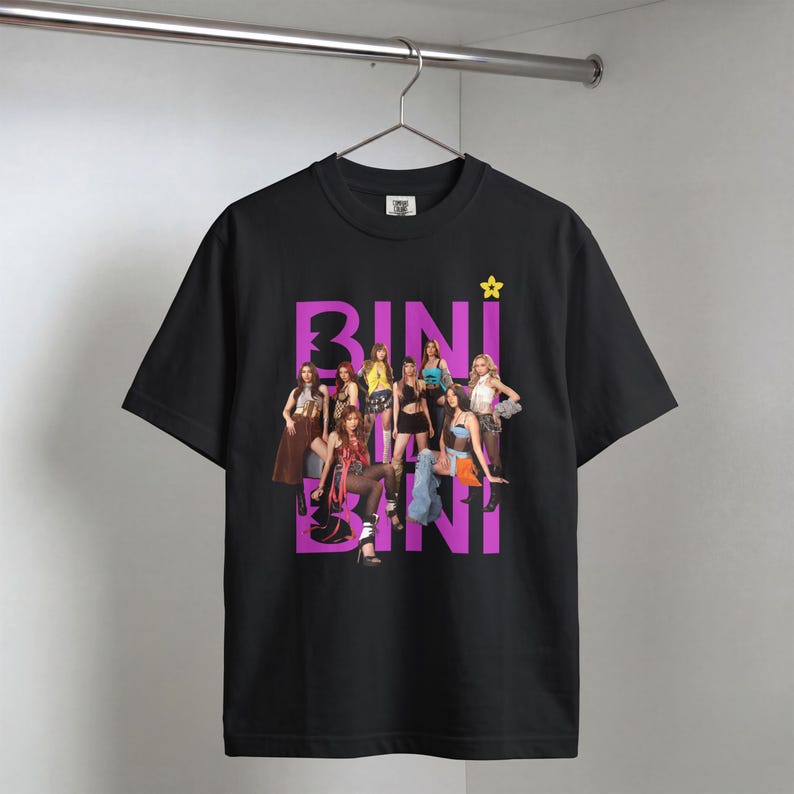 May include: A black short-sleeved t-shirt featuring a graphic print of a group of eight people posing. The text BINI is printed in large purple letters behind the group, with a small yellow star icon above the text. The shirt hangs on a hanger.