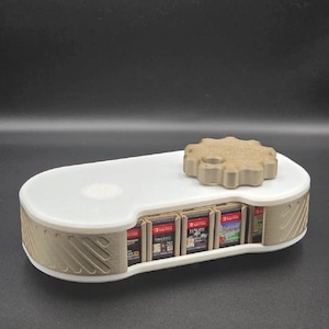 Puede incluir: Un soporte para cartuchos de juego impreso en 3D, blanco y beige. El soporte tiene una base rectangular con un diseño curvo y una tapa beige en forma de engranaje. Varios cartuchos de juego son visibles dentro.