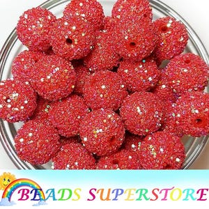 May include: A collection of red, glitter-covered beads in a clear glass bowl. The beads are round with a hole through the center, and the surface is covered in small, sparkling glitter. The image is well-lit, highlighting the texture and color of the beads.