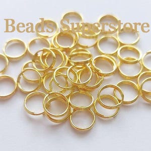 May include: A collection of gold-coloured metal jump rings for jewellery making, scattered across a plain surface. The text Beads Superstore is overlaid in a light brown font across the top portion of the image, centred above the metal rings.