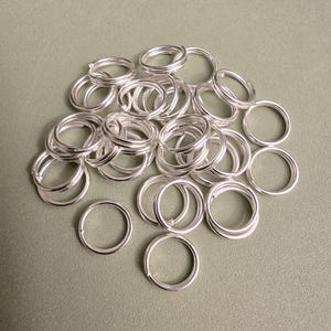 May include: A collection of silver-colored metal split rings arranged in a loose pile. Each circular ring features a double-loop design, creating a coiled appearance with a smooth, reflective finish. The rings are uniform in shape and size.