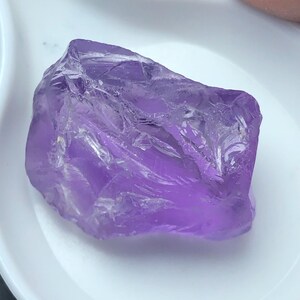 Könnte beinhalten: Ein roher, durchscheinender violetter Amethyst-Edelstein mit unregelmäßiger, gezackter Form und kristalliner Textur. Der Stein zeigt verschiedene Schattierungen von Violett und Dunkellila auf seiner facettierten, natürlichen Oberfläche.