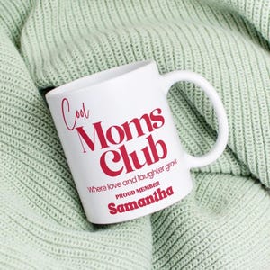 Puede incluir: Taza de cerámica blanca con el texto "Cool Moms Club" en rojo. La taza también incluye las frases "Where love and laughter grow" y "Proud Member Samantha". La taza está sobre una manta de punto verde claro.