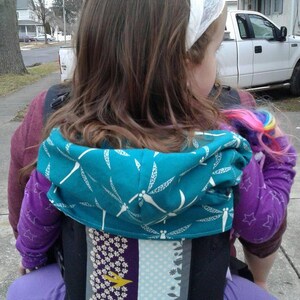 May include: A teal baby carrier with a dragonfly pattern on the hood. The carrier has a black base with decorative panels in purple, light blue, and gray. The child is wearing a purple shirt with star patterns.