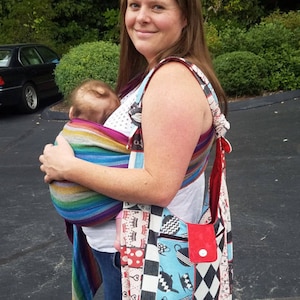 Può includere: Una donna indossa un marsupio a righe blu e bianche con un bambino che dorme dentro. Porta anche una borsa a tracolla a motivi bianco e nero con una tasca rossa.