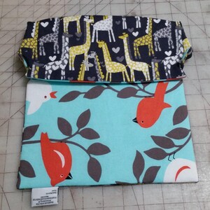 May include: A folded fabric pouch with a giraffe pattern on the top flap and a bird and branch pattern on the main body. The giraffe pattern features yellow, gray, and white giraffes on a navy blue background. The bird pattern has red birds and gray branches on a light blue background.