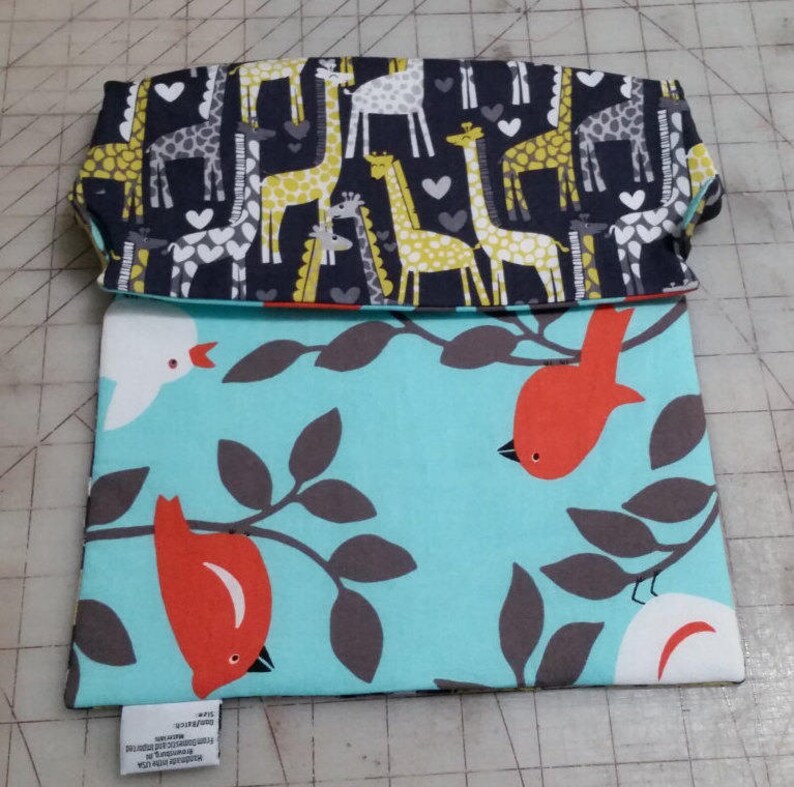 May include: A folded fabric pouch with a giraffe pattern on the top flap and a bird and branch pattern on the main body. The giraffe pattern features yellow, gray, and white giraffes on a navy blue background. The bird pattern has red birds and gray branches on a light blue background.