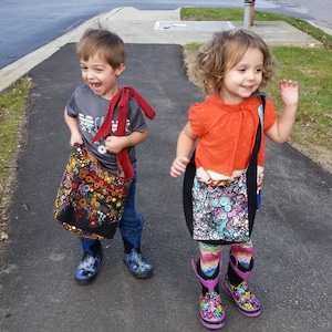 Może przedstawiać: Dwoje dzieci, chłopiec i dziewczynka, noszą kolorowe, wzorzyste torby. Chłopiec ma na sobie szarą koszulę z nadrukiem graficznym i niebieskie dżinsy. Dziewczynka ma na sobie jasno pomarańczową koszulę i kolorowe, pasiasty rajstopy. Oboje dzieci uśmiechają się i patrzą w obiektyw.