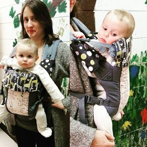 May include: A baby carrier with a black and white polka dot pattern and a giraffe print. The carrier is gray and pink with adjustable straps and buckles. The baby is wearing a white long-sleeved shirt.