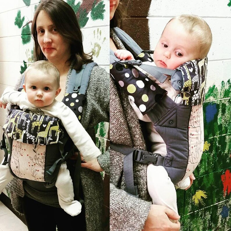 May include: A baby carrier with a black and white polka dot pattern and a giraffe print. The carrier is gray and pink with adjustable straps and buckles. The baby is wearing a white long-sleeved shirt.
