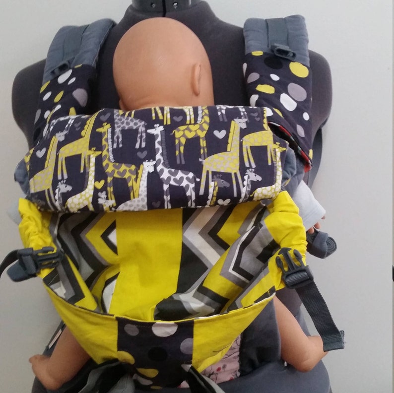 May include: A baby carrier with a giraffe pattern in yellow and white on a navy blue background. The carrier has gray straps and a yellow and gray geometric patterned front. The carrier is designed to hold a baby.