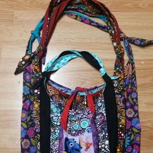 Può includere: Una borsa a tracolla in tessuto multicolore con fodera in tessuto nero. La borsa ha una tracolla lunga e una piccola tasca sul davanti con un pesce dei cartoni animati.