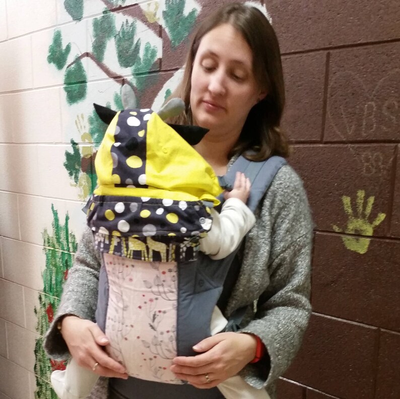 May include: A baby carrier with a yellow and navy blue cow-themed hood. The carrier has a pink and gray floral patterned panel. The baby is wearing a white onesie. The person holding the baby is wearing a gray sweater.