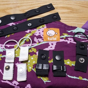 以下が含まれることがあります： チュラベビーキャリア用の黒と白のストラップ、バックル、リングのセット。ストラップには黒いボタンが付いており、バックルは黒いプラスチック製です。リングは白いプラスチック製です。ストラップは、オレンジ色の「Tula」という文字が書かれた、紫色の花柄の生地の上に置かれています。