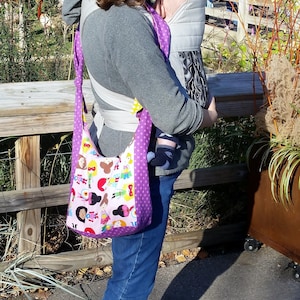 May include: A woman wearing a purple and white patterned sling bag with a baby carrier. The bag has a colorful cartoon design of girls wearing different outfits.