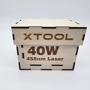 Puede incluir: Una caja de madera apilada con el texto "XTOOL" en la parte superior y "40W 455nm Laser" en la inferior. La caja es de madera clara con detalles marrón oscuro en las esquinas y bordes. Para láser.