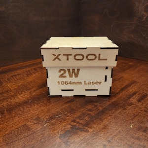 Pode incluir: Uma caixa de madeira clara com o texto "XTOOL" na parte superior e "2W 1064nm Laser" na frente. A caixa está sobre uma superfície de madeira.