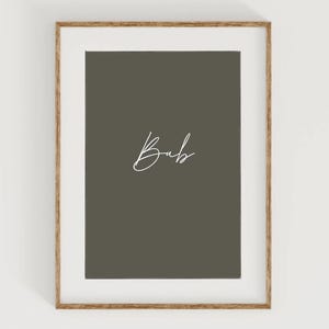 May include: A framed print featuring the word "Bub" in elegant white script on a muted olive-green background. The print is set within a white mat and a natural wood-toned frame, creating a minimalist aesthetic.