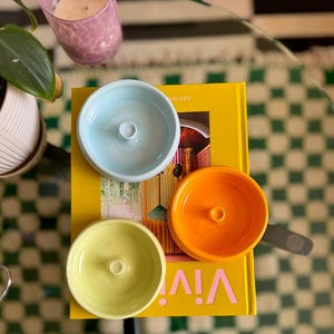 May include: Four ceramic bowls in different colors: light blue, yellow, orange, and white. The bowls are stacked on top of a yellow book with the word "VIV!" printed on the cover. The bowls have a small hole in the center.