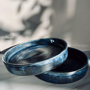 May include: Two handmade, ceramic, oval-shaped dishes. The dishes are a deep blue with a glossy finish and a lighter blue rim. The dishes are stacked on top of each other, with one slightly overlapping the other.